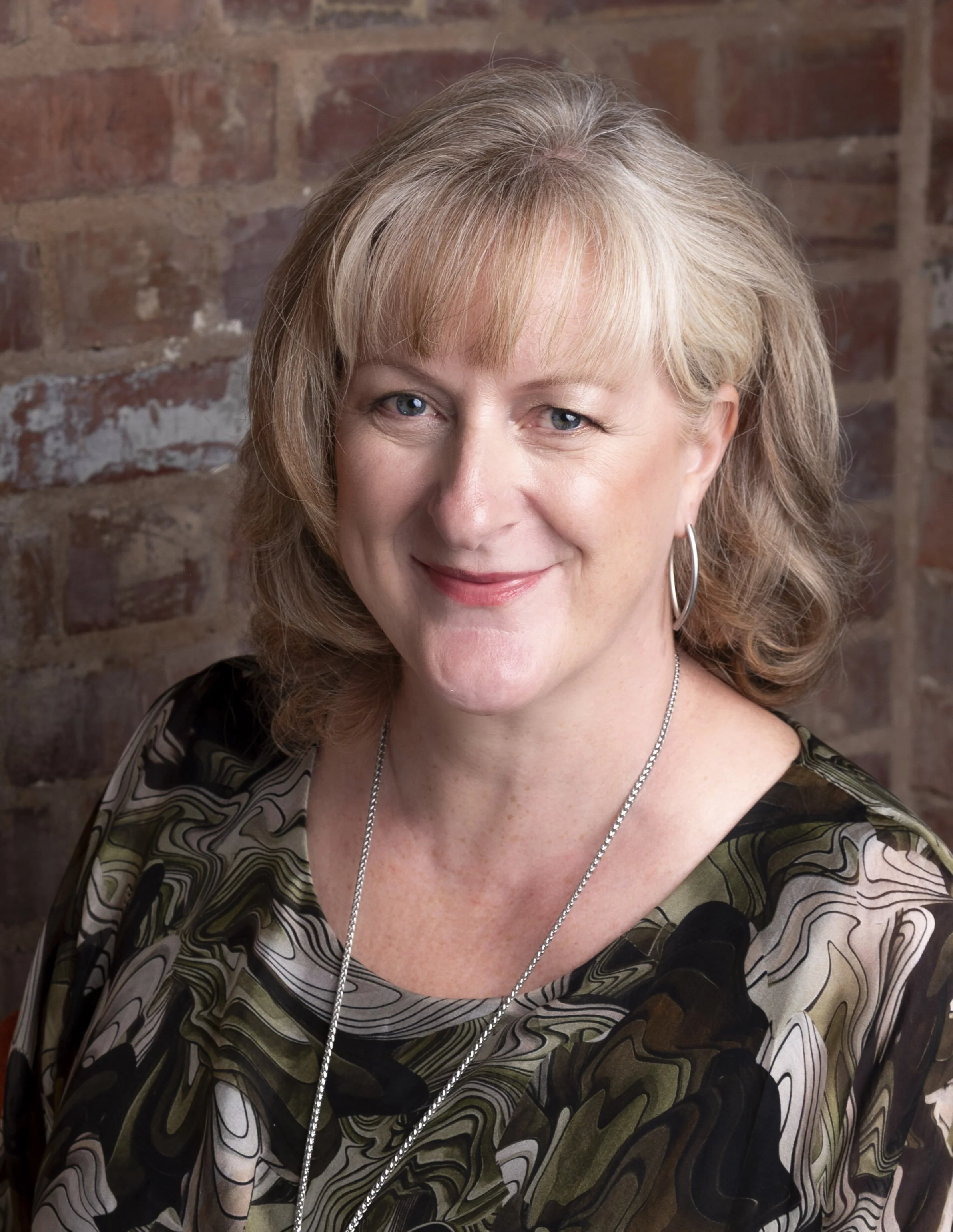 Headshot of a smiling woman with a blonde bob and a patterned top in front of a brick wall