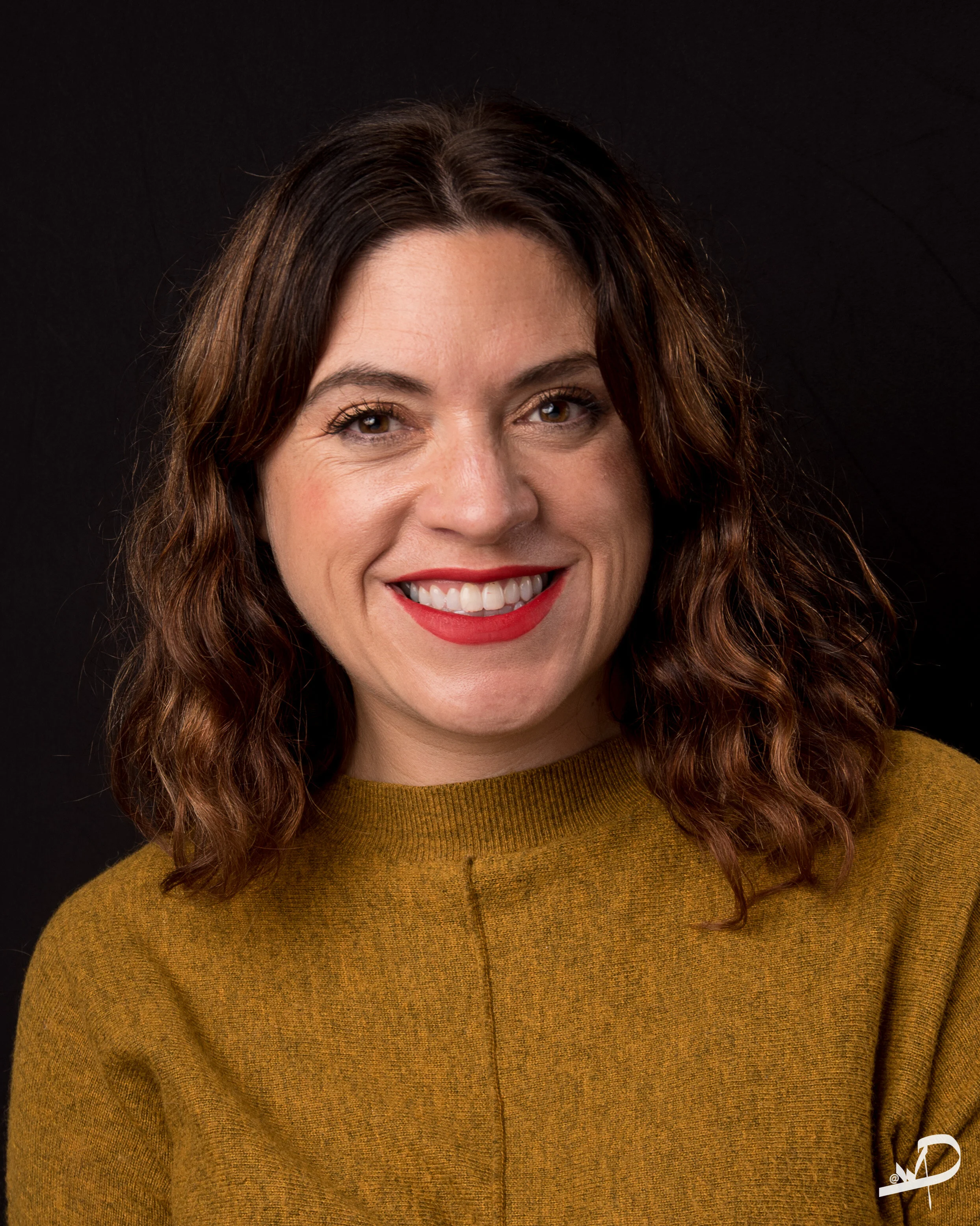 A woman with shoulder-length brown curly hair, wearing a mustard-colored sweater, smiling against a black background.