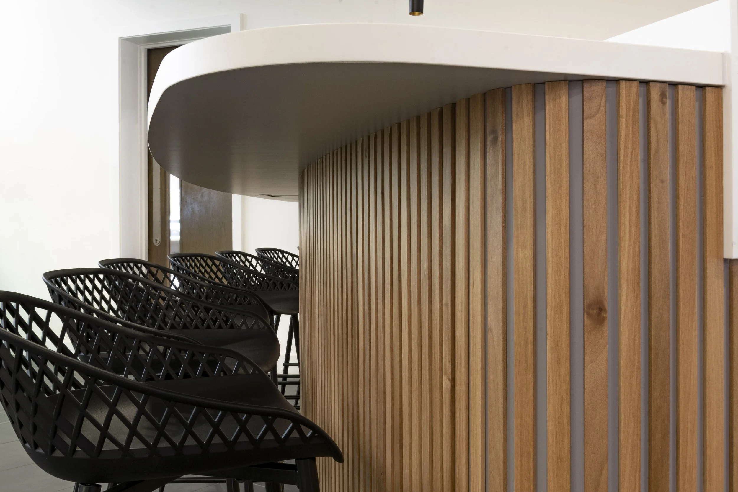 Curved timber-slatted counter with black mesh stools beneath a white canopy