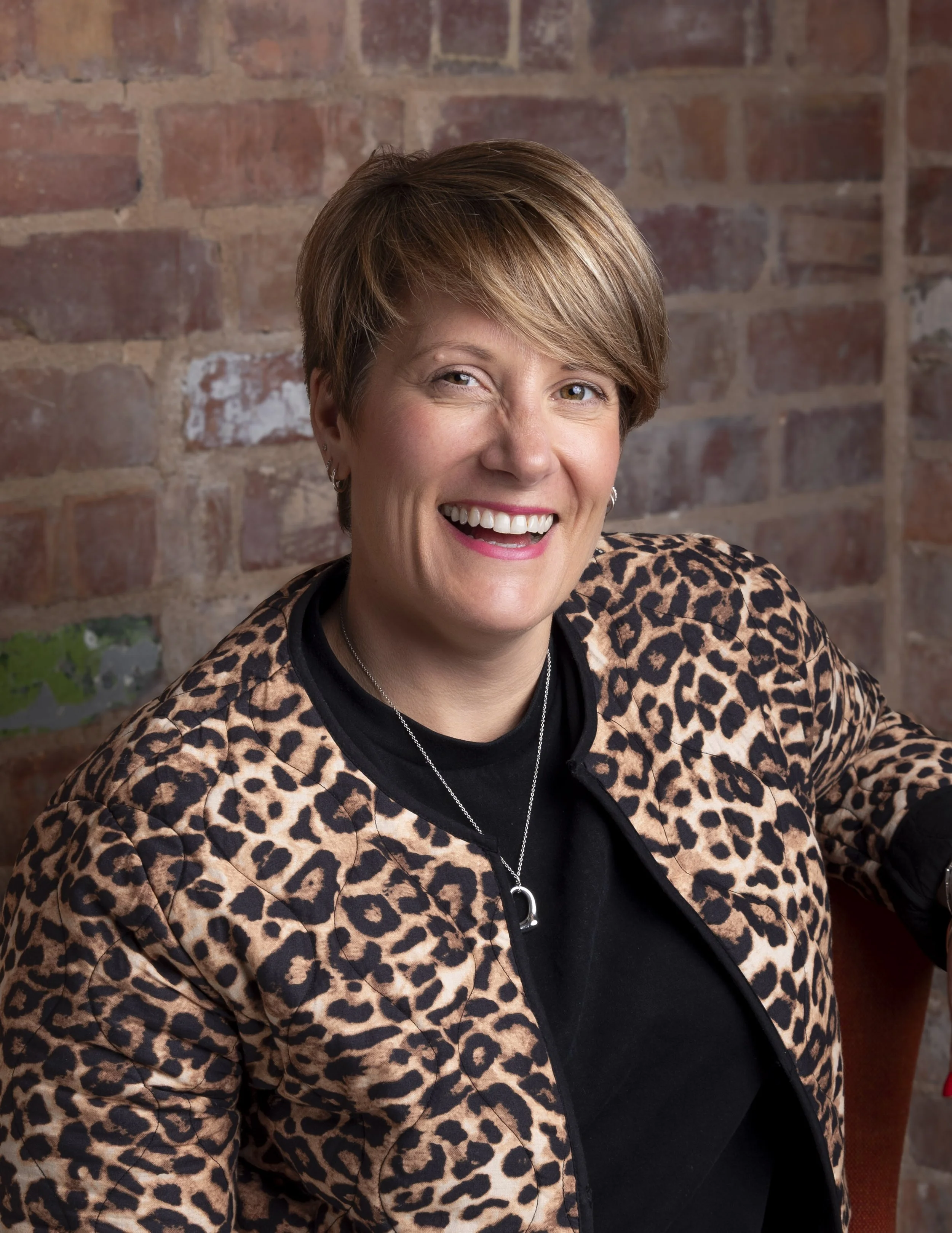 Laughing woman with short hair in a leopard print jacket in front of a brick wall