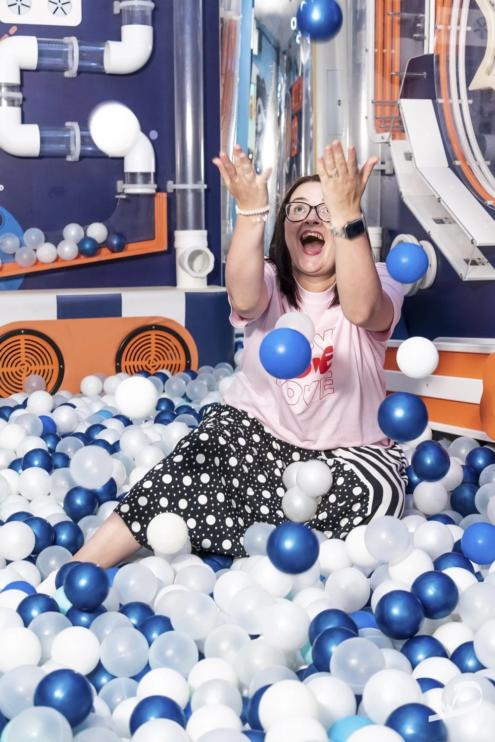 A woman in a pink shirt and polka dot pants sitting in a ball pit with white, blue, and teal balls, smiling and tossing balls in the air, in an indoor play area.