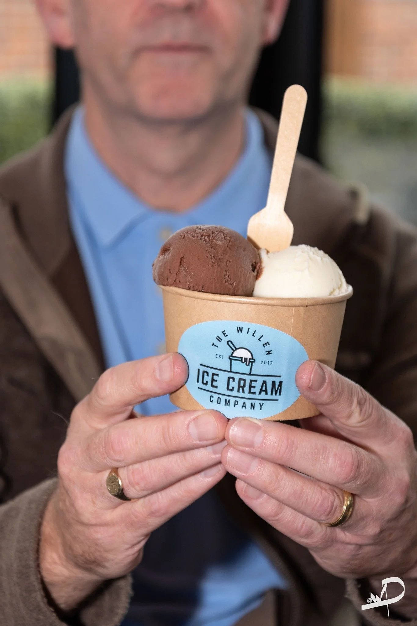 A person holding a cup of ice cream with two scoops, one chocolate and one vanilla, with a small wooden spoon, in front of a blurred background.