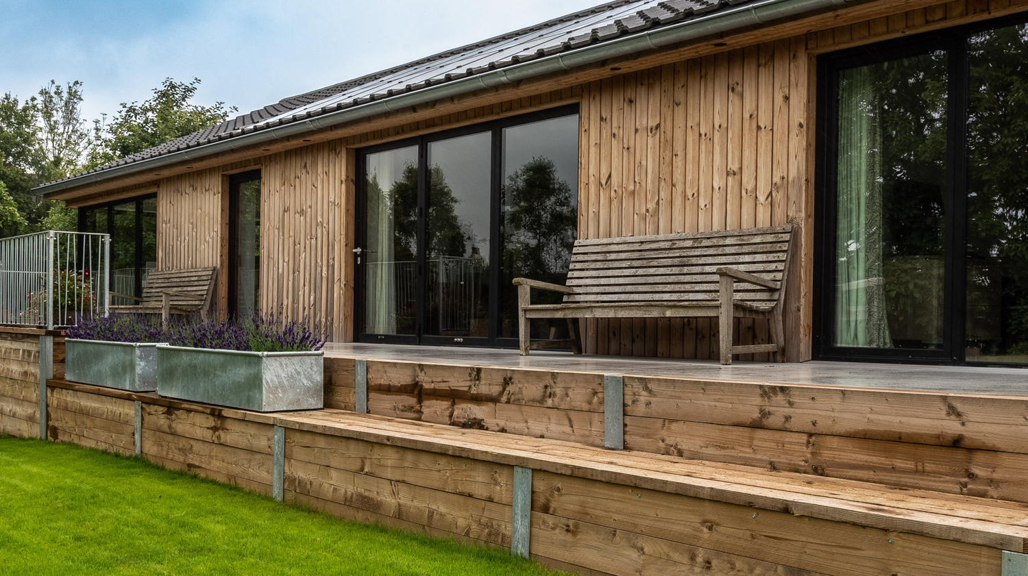 Wooden house with large glass sliding doors and a balcony with benches and plant boxes, overlooking a green lawn.