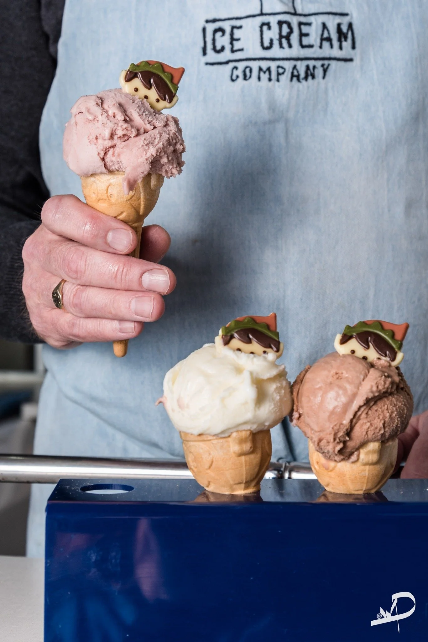Person holding an ice cream cone with pink ice cream and a cookie decoration, two additional ice cream cones with vanilla and chocolate flavors and cookie decorations on a blue stand.