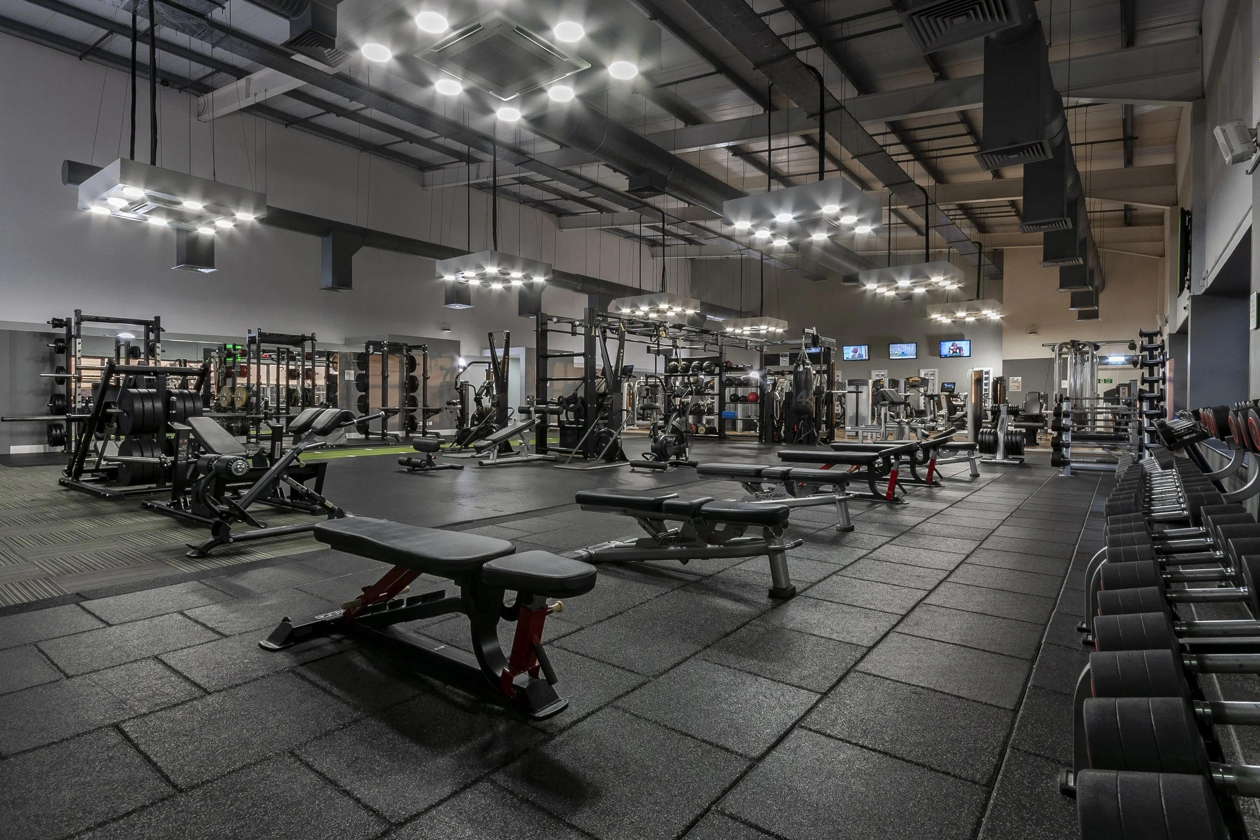 Wide view of a gym floor with benches, racks and overhead spotlights