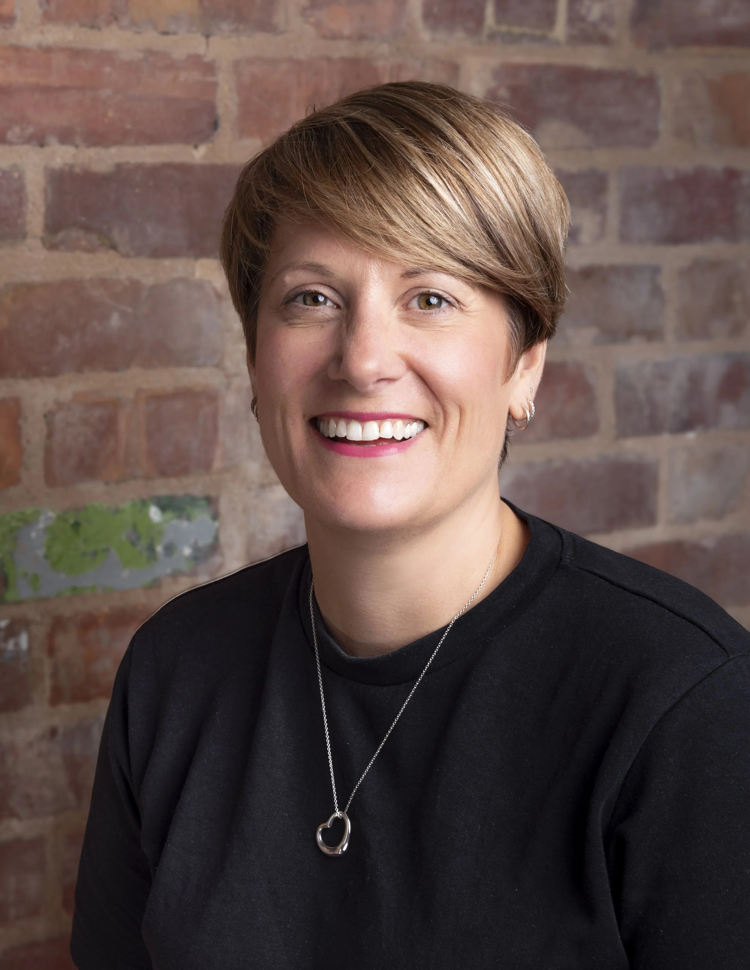 Headshot of a smiling woman with short styled hair and a black top in front of a brick wall