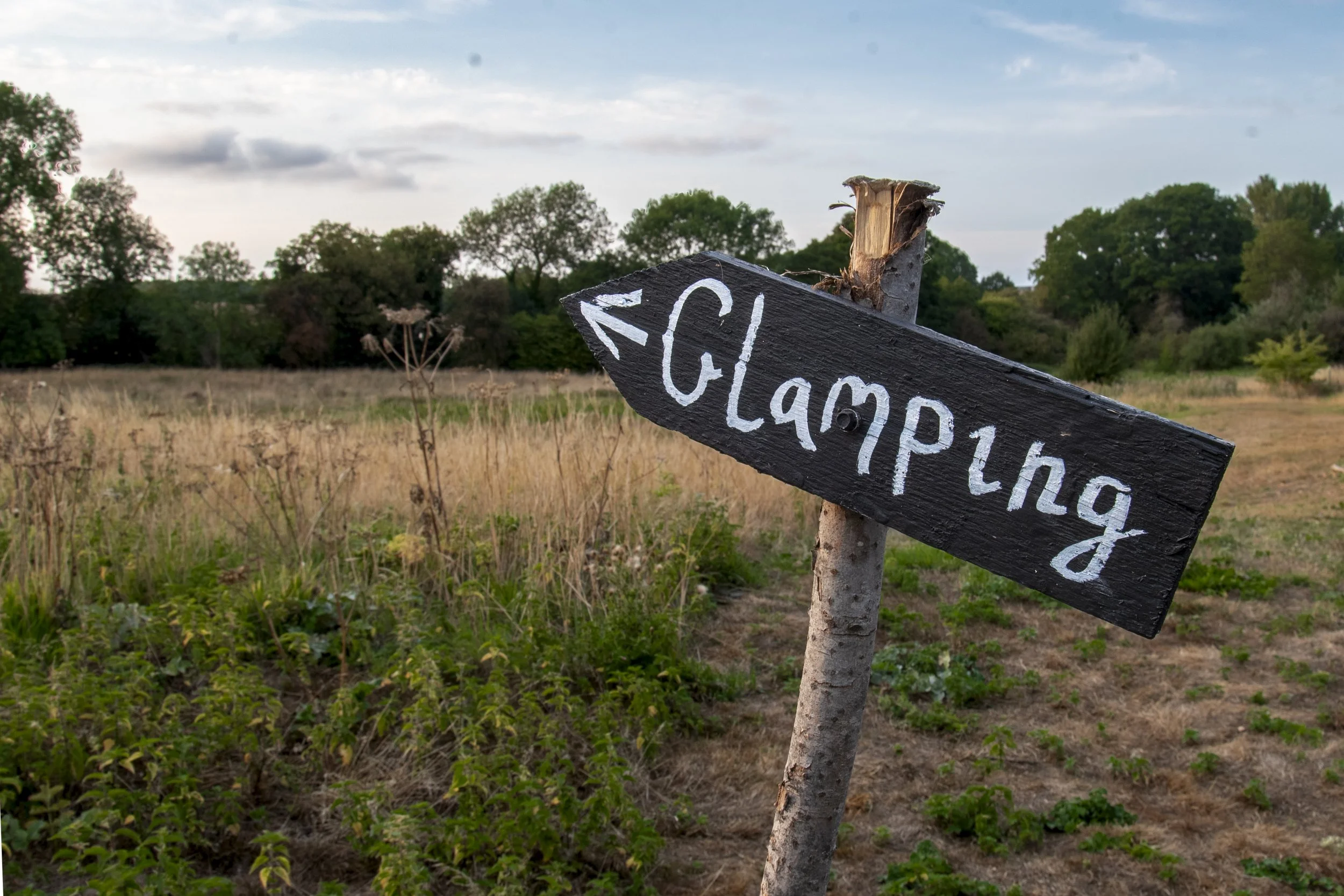 A black wooden sign with white lettering reading 'Clamping' and an arrow pointing to the left, posted on a wooden stake in a rural field with trees and dried grass.