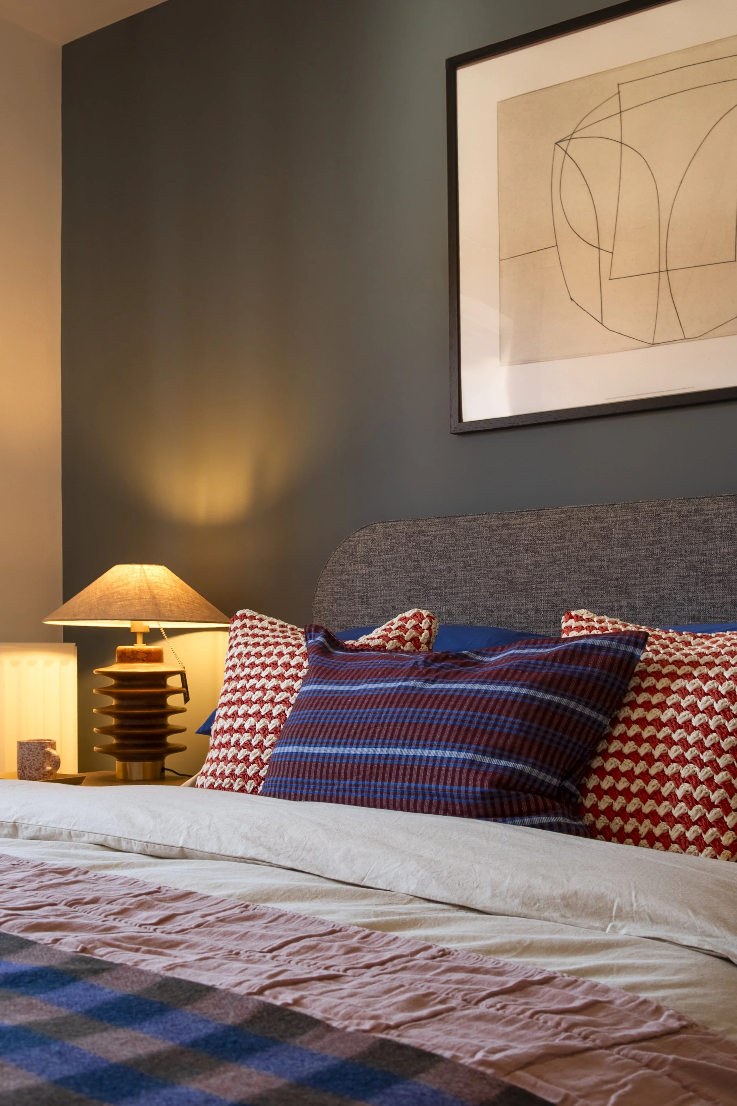 Close-up of a bed with a gray upholstered headboard, patterned pillows, and a blanket, beside a warm lit table lamp and a decorated wall.
