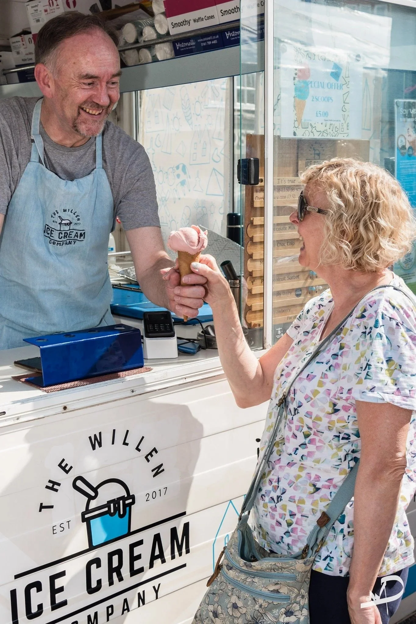 A woman with curly blonde hair and sunglasses is accepting a pink ice cream cone handed by a smiling ice cream vendor at a booth. The booth has a sign that says 'The Willen Ice Cream Company' established 2017.