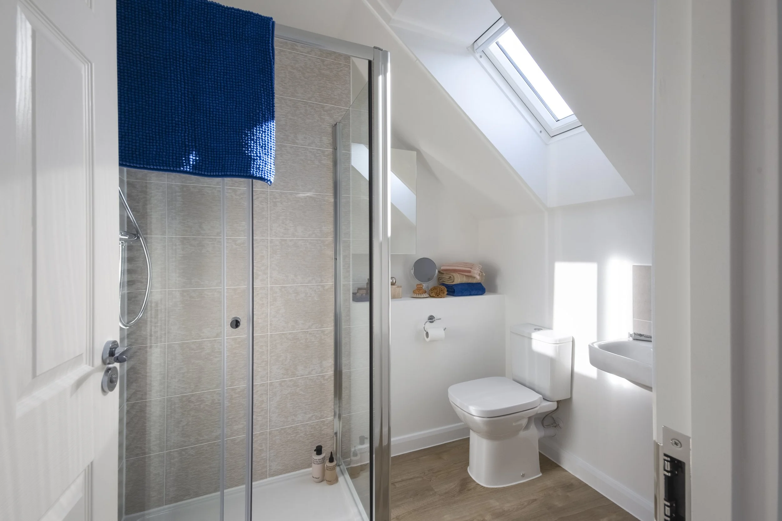 A modern bathroom with a glass shower stall, a toilet, a small sink, and a skylight window, with towels and toiletries arranged on a shelf.