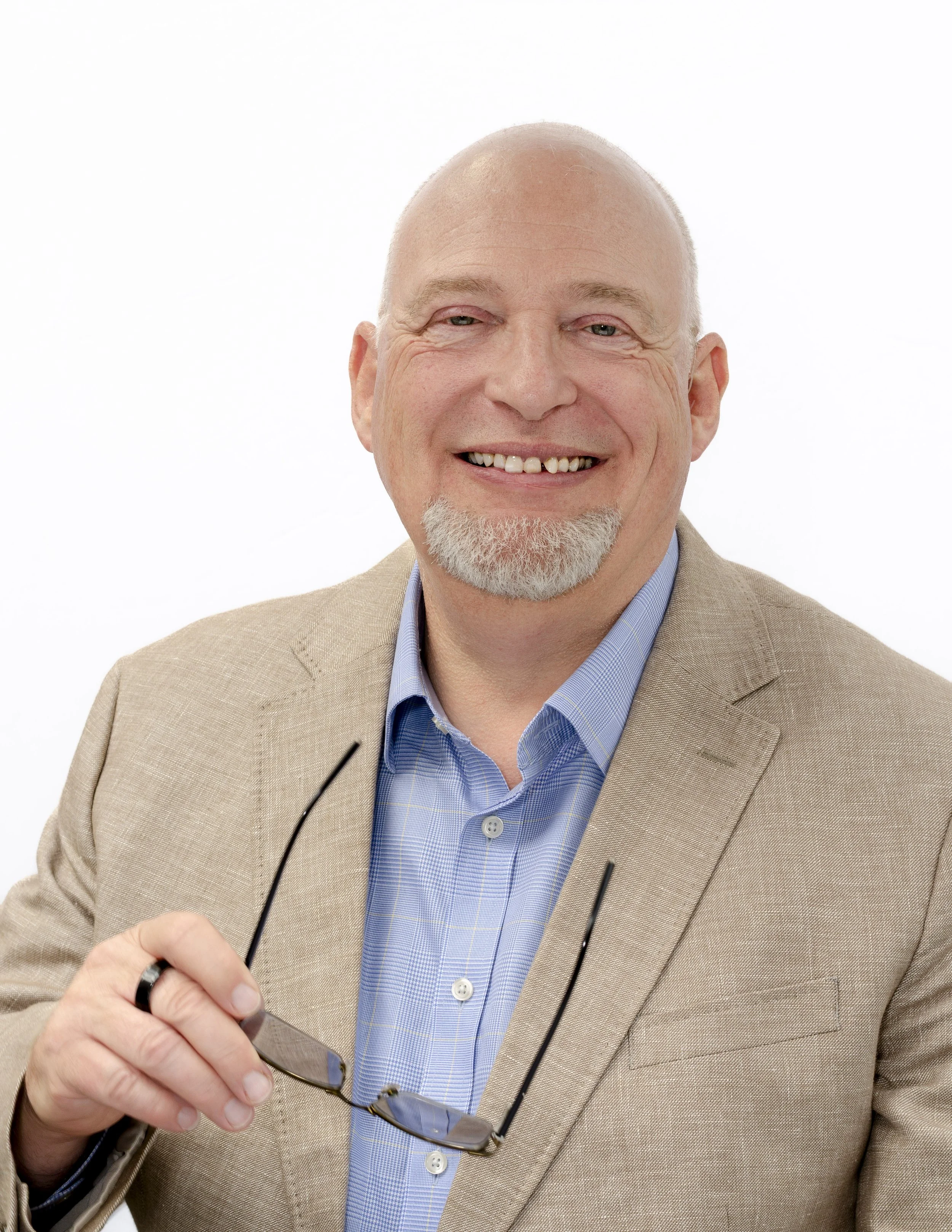 Mark Spence smiling in a beige blazer, holding his glasses, on a white background