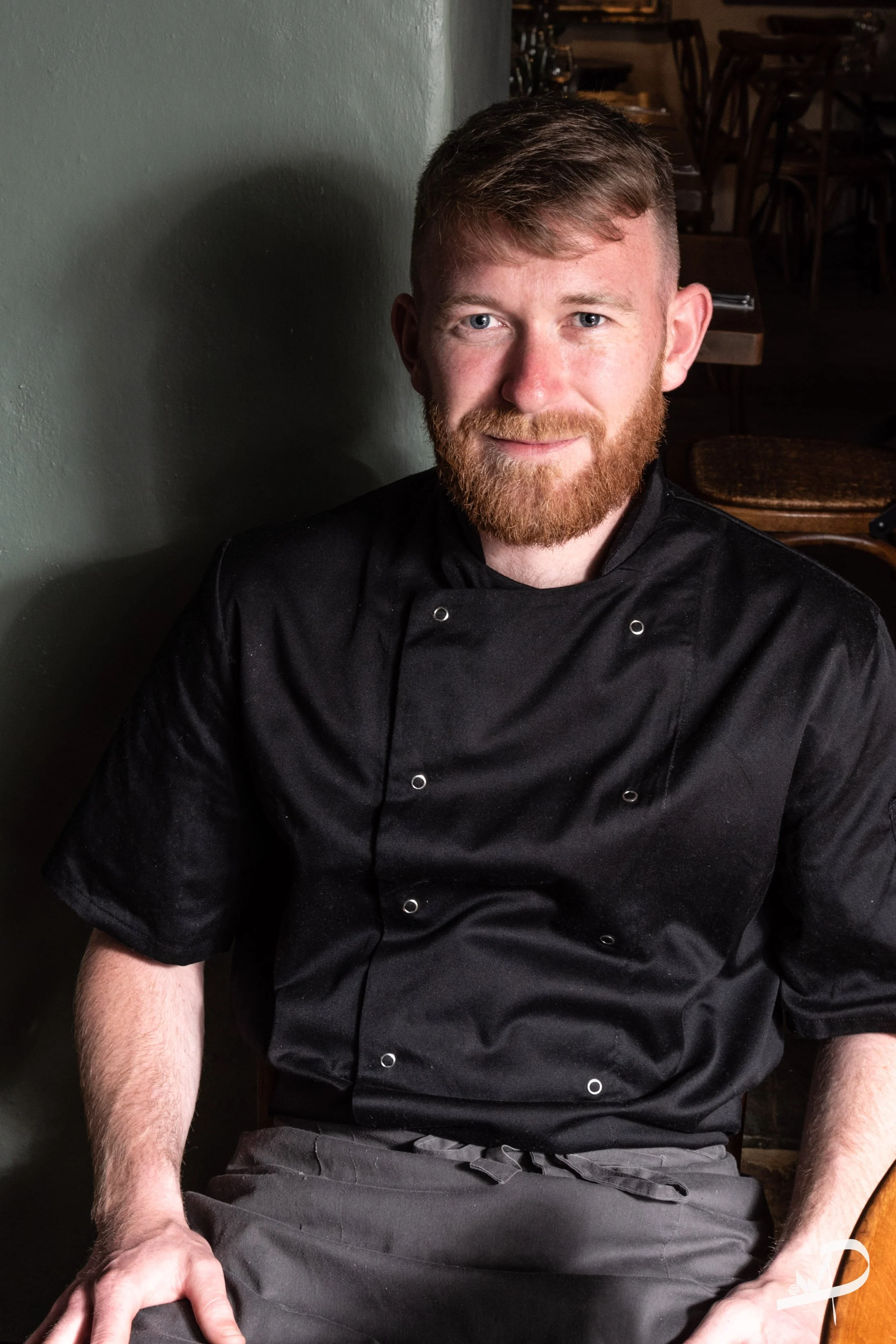 A male chef with red hair and beard wearing a black chef coat sitting in a restaurant or kitchen area.