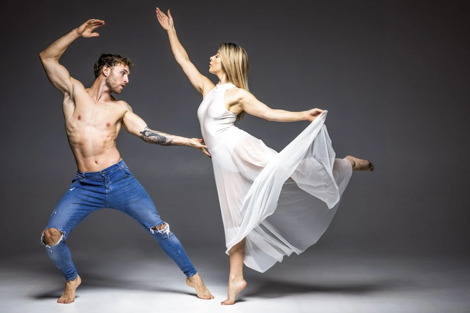 A shirtless man in ripped jeans and a woman in a flowing white dress dance together against a gray background, with the woman balancing on one leg and the man reaching towards her.