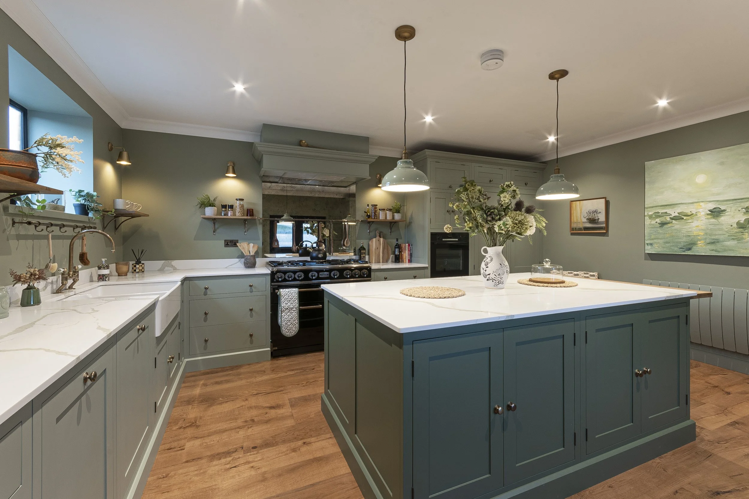 A spacious kitchen with green cabinets, a large white marble island, and wooden flooring. There are pendant lights, open shelves with decor, a black stove, and a vase with flowers on the island. The walls are painted green and there's artwork on the wall.