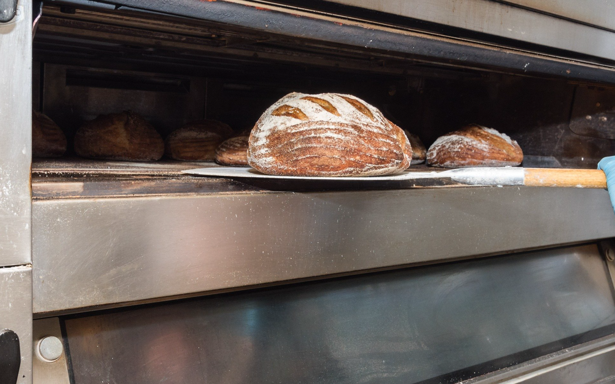 Loaf of round artisan bread with scoring on top, placed on a baking sheet inside a commercial oven.