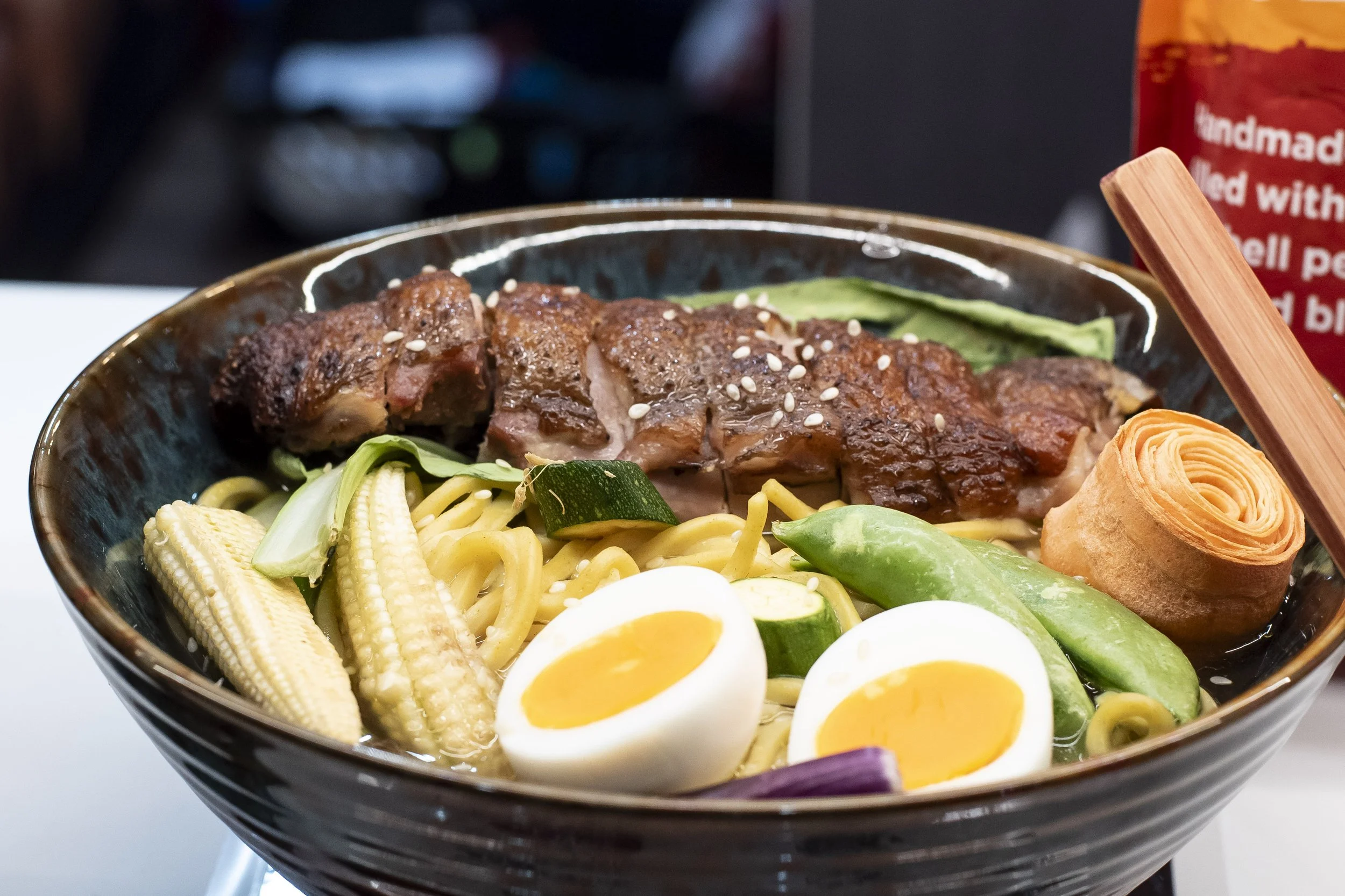 Bowl of noodles topped with sliced steak, baby corn, mange tout and a halved soft-boiled egg