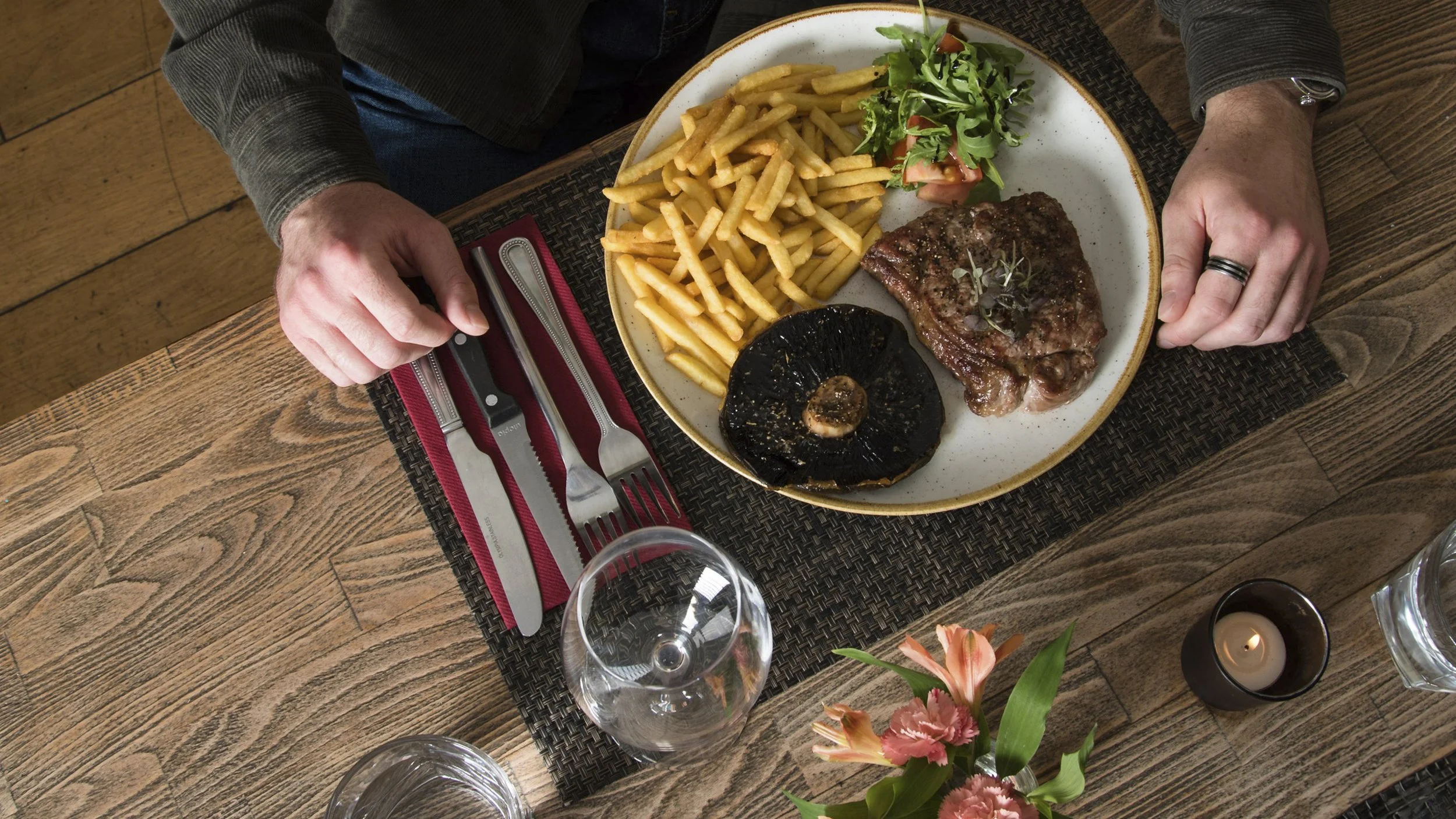 A person sitting at a wooden table with a plate of steak, French fries, a grilled mushroom, and a small salad. The table has a red napkin with utensils, a wine glass, a flower vase with pink flowers, and a lit candle.