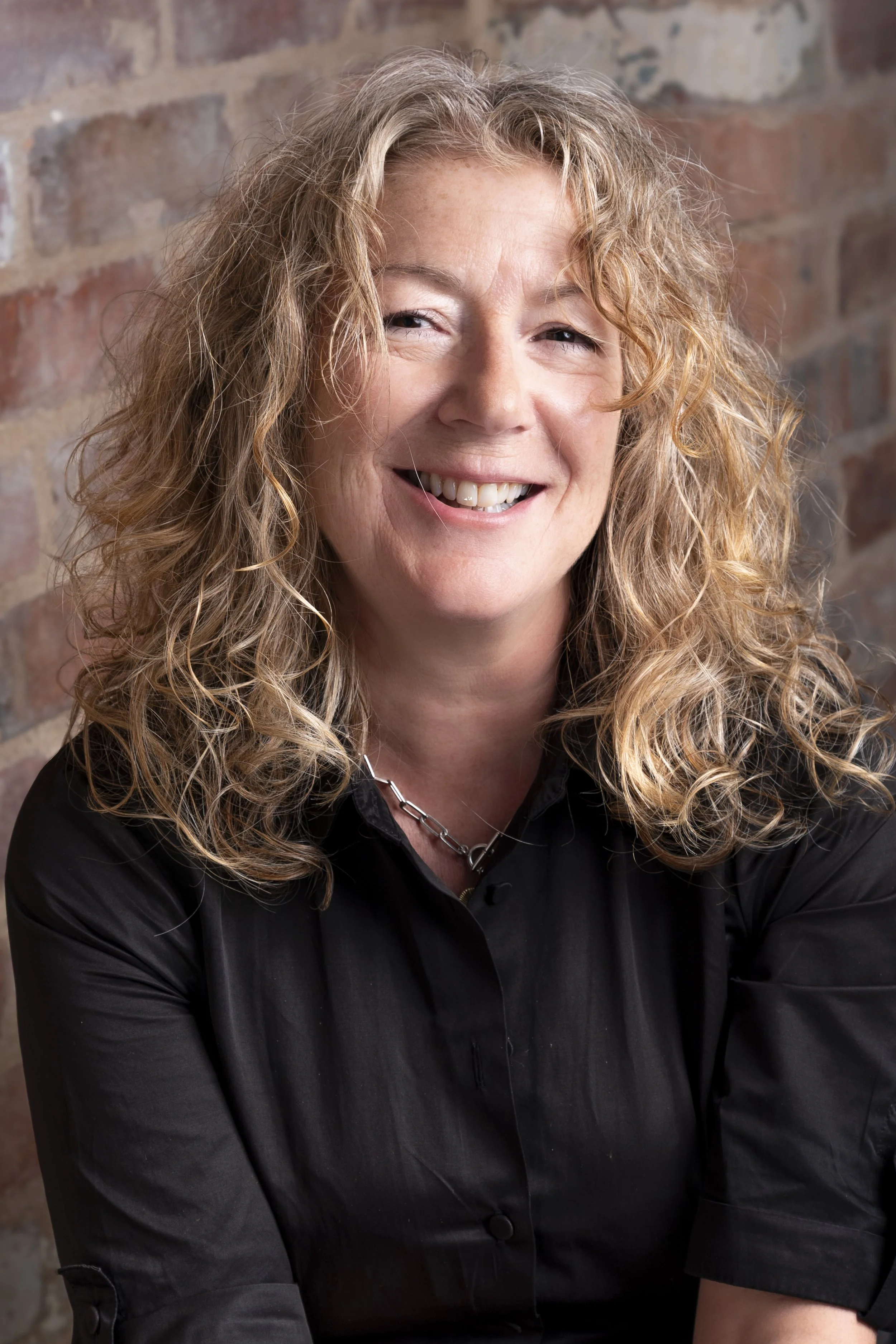 Close-up headshot of a smiling woman with curly blonde hair and a black top in front of a brick wall