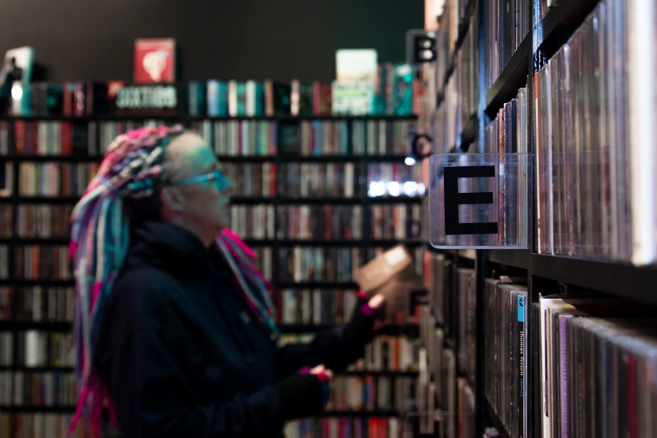 A person with colorful braids and glasses browsing books in a dimly lit library or bookstore.