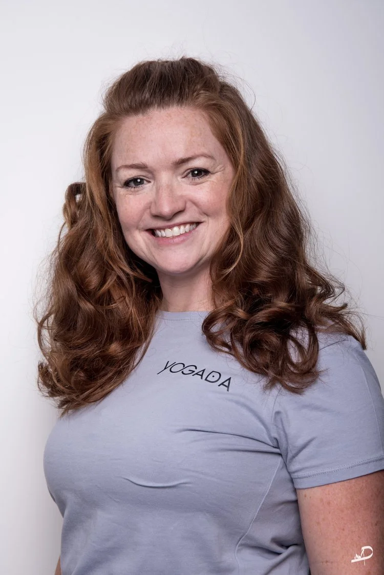 A woman with long, curly red hair smiling at the camera, wearing a light gray T-shirt with the word 'YOGADA' printed on it, standing against a plain white background.
