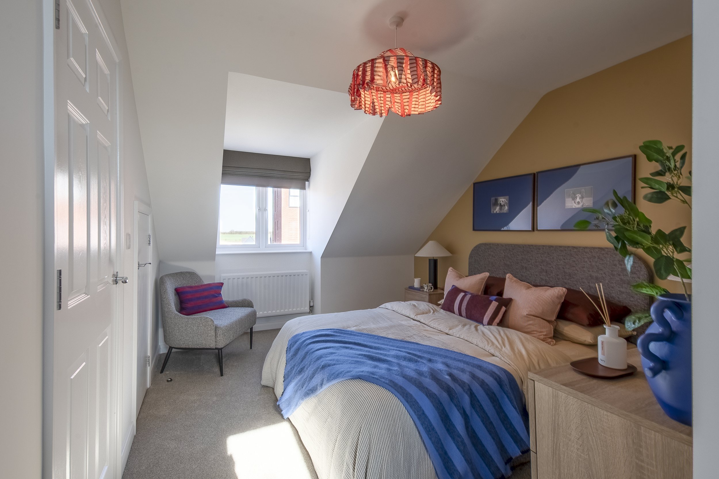 A cozy bedroom with a sloped ceiling, a bed with pillows and a blue striped blanket, a lamp on the bedside table, artwork above the bed, a window letting in natural light, an armchair near the window, and a decorative plant in a blue vase.
