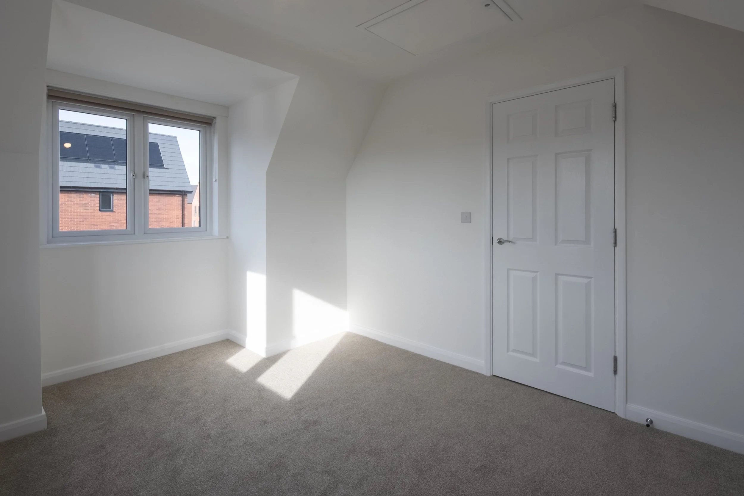 Empty room with beige carpet, white walls, a white door, and a window showing neighboring houses.