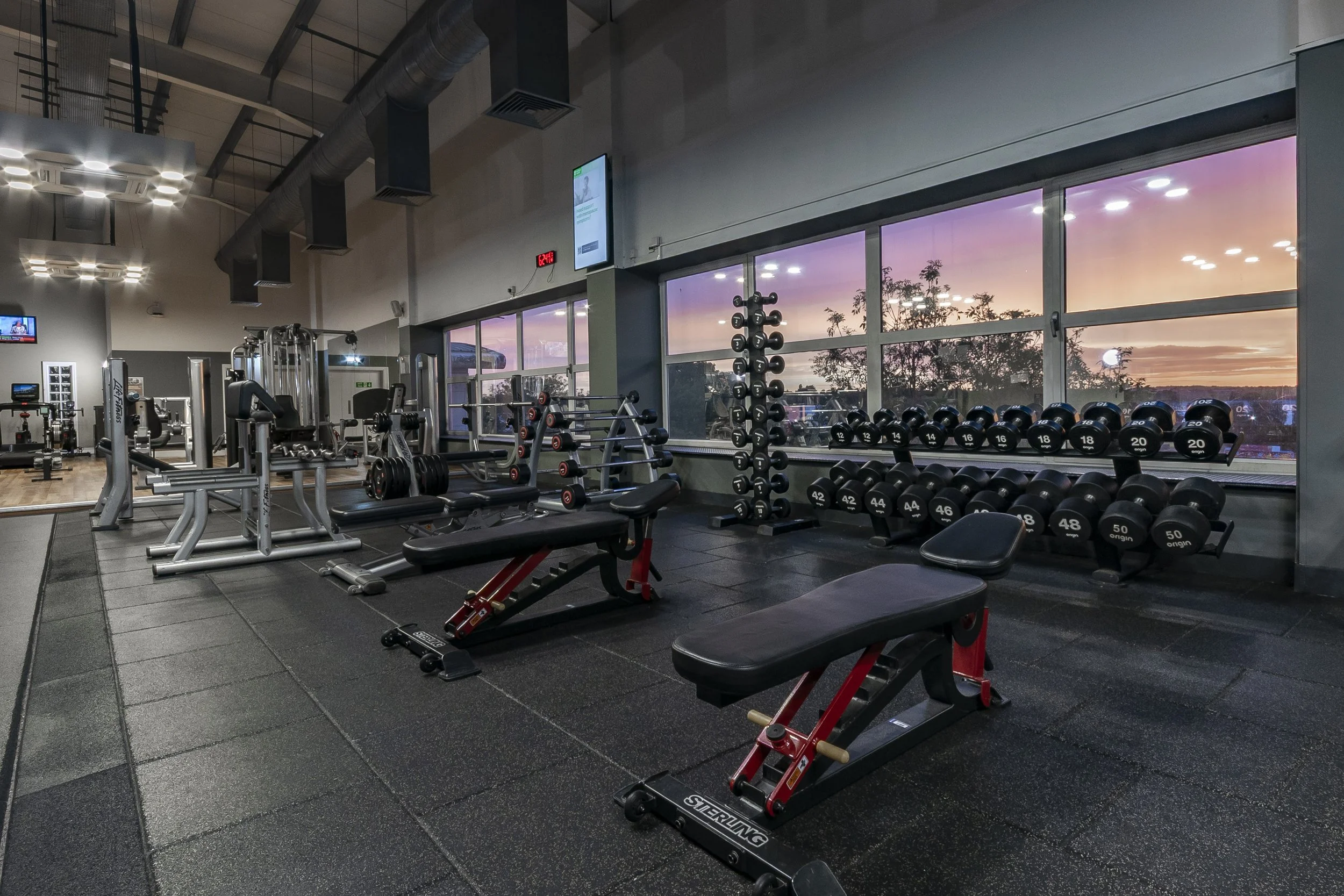 Gym floor with benches and dumbbell rack lit by pink sunrise through large windows