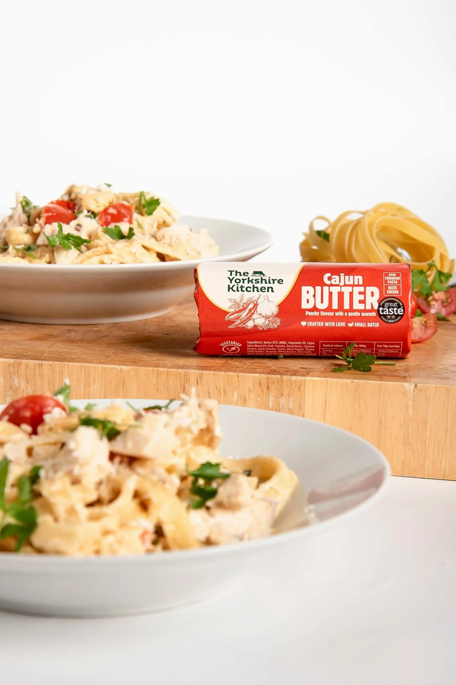 A block of Cajun butter from The Yorkshire Kitchen placed on a wooden surface, with a plate of creamy pasta with cherry tomatoes and herbs and a bowl of pasta in the background.