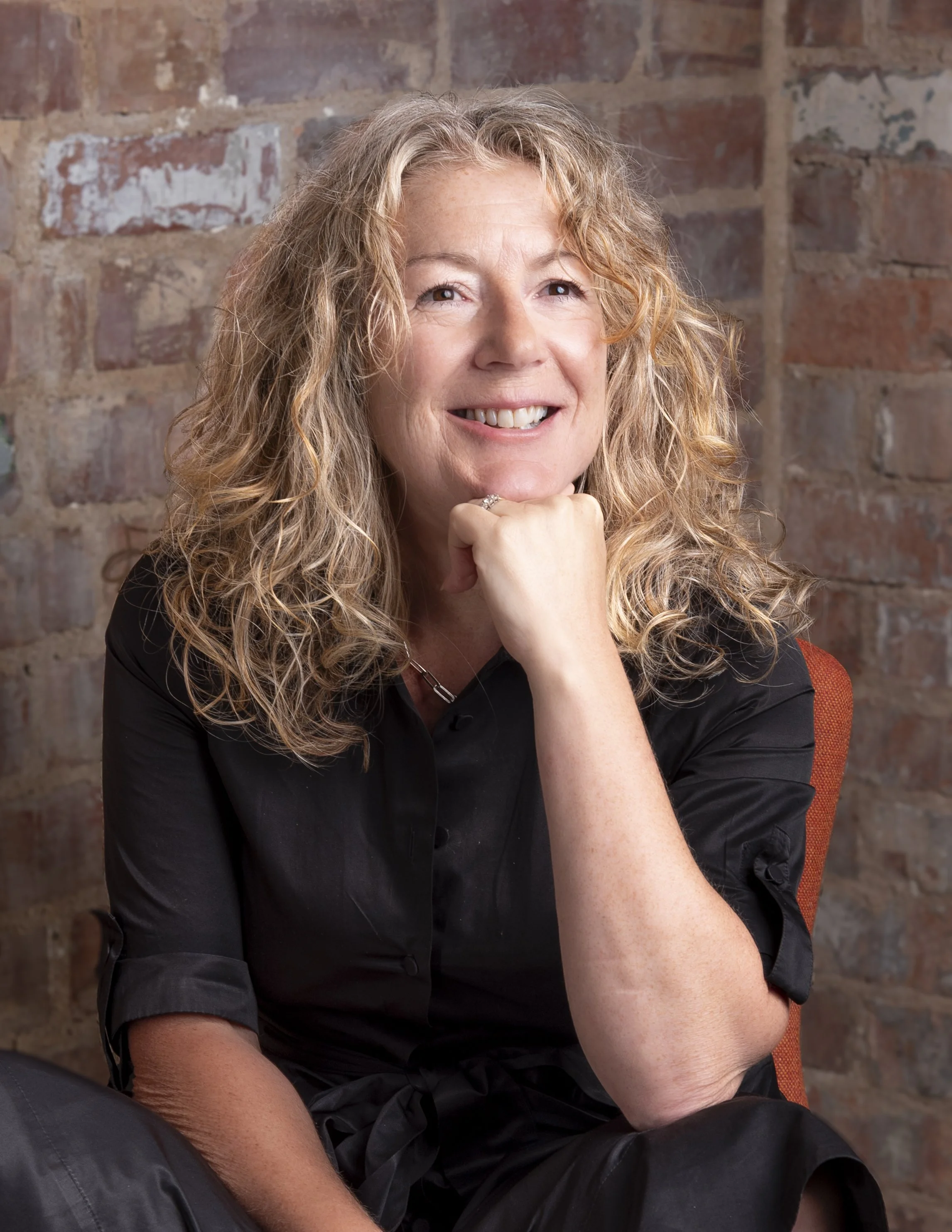 Woman with curly blonde hair in a black shirt resting her chin on her hand in front of a brick wall