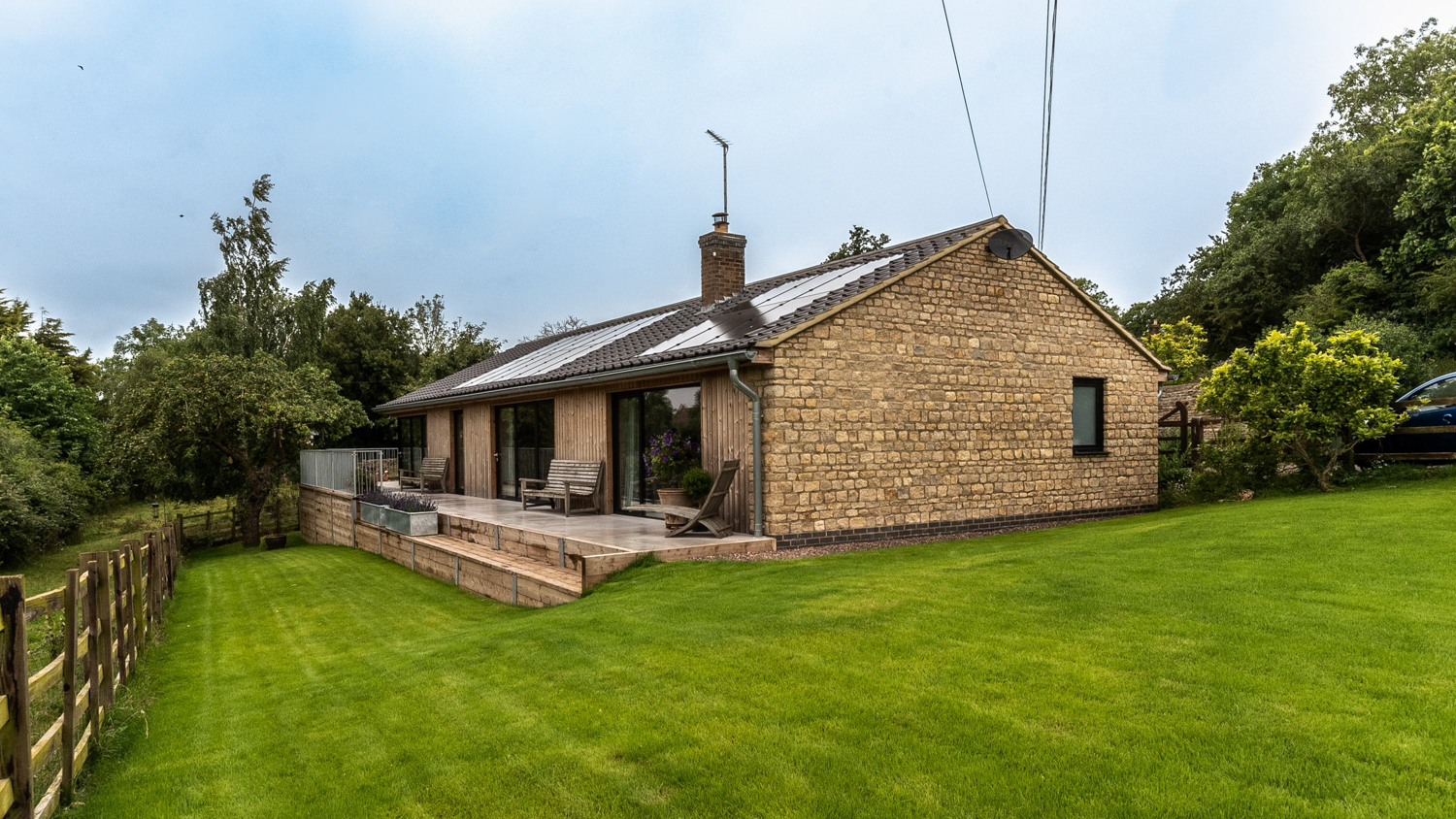 Brick house with a sloped roof, solar panels, a chimney, and a nearby parked car, surrounded by lush green grass and trees.