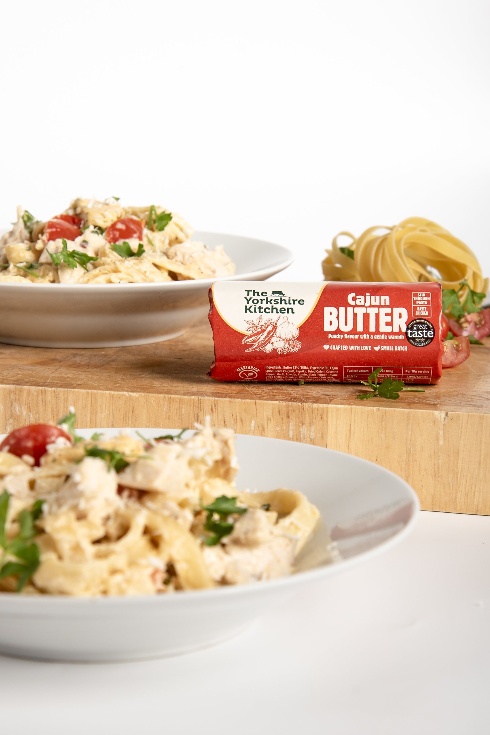 A white bowl of creamy chicken and pasta salad with cherry tomatoes and parsley, a package of The Yorkshire Kitchen Cajun Butter on a wooden surface, a bunch of cooked pasta in the background, and a similar pasta dish in the foreground.