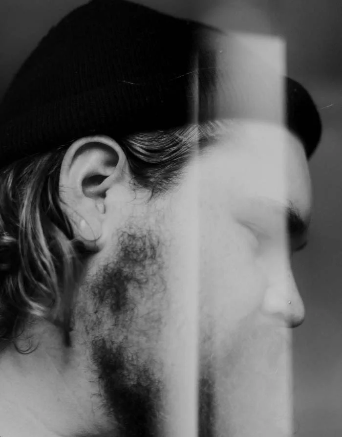 A black and white photo of a man with a beard, wearing a knit cap, seen in profile from his left side, with a reflection of a window or bright light in the background.