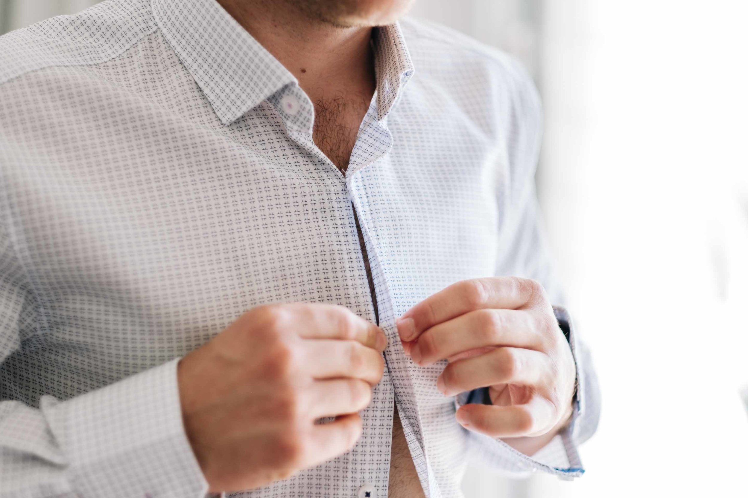 a closeup of the groom buttoning up his shirt
