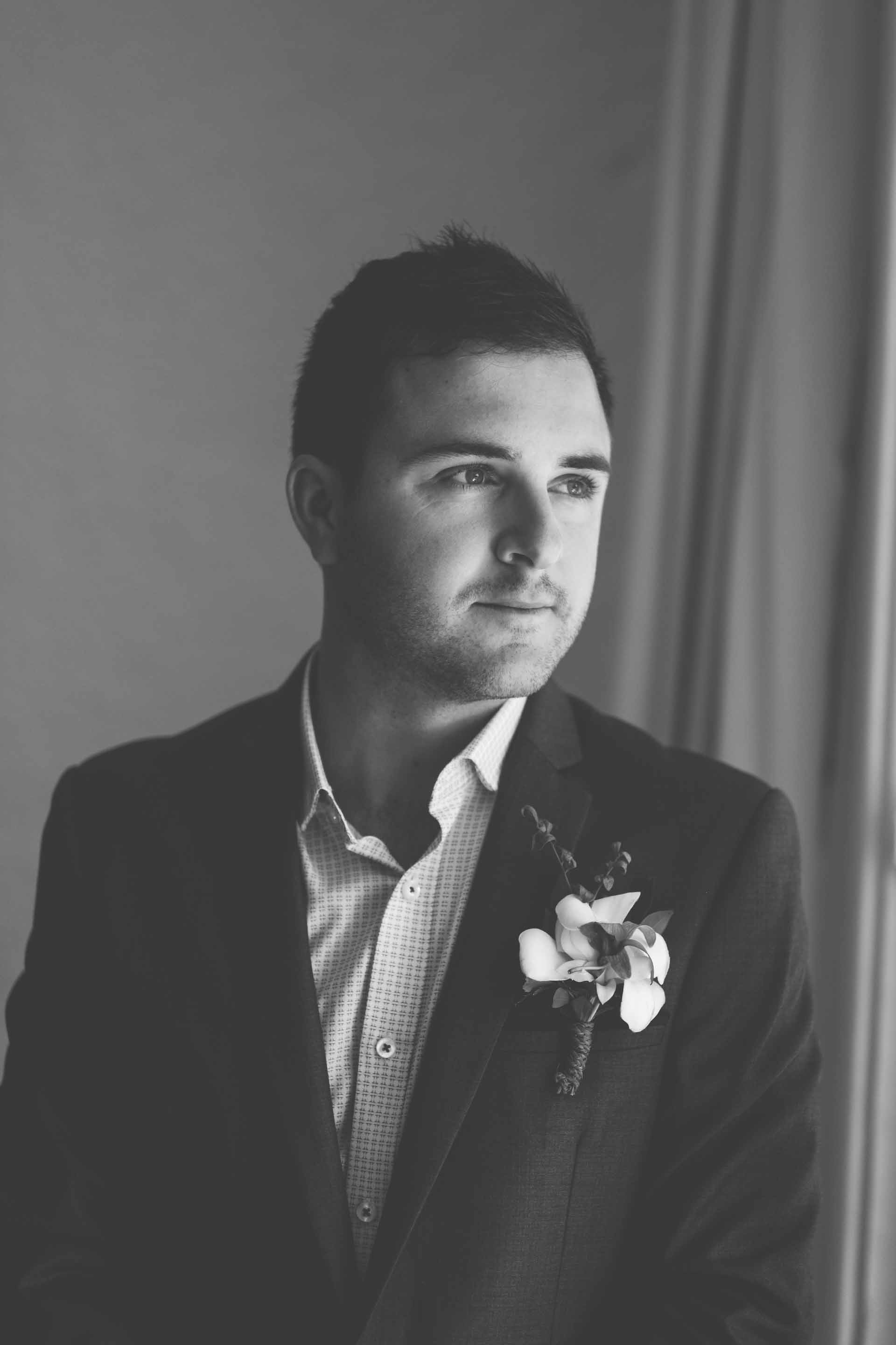 a black and white portrait of the groom looking out the window wearing his wedding suite with boutonnière
