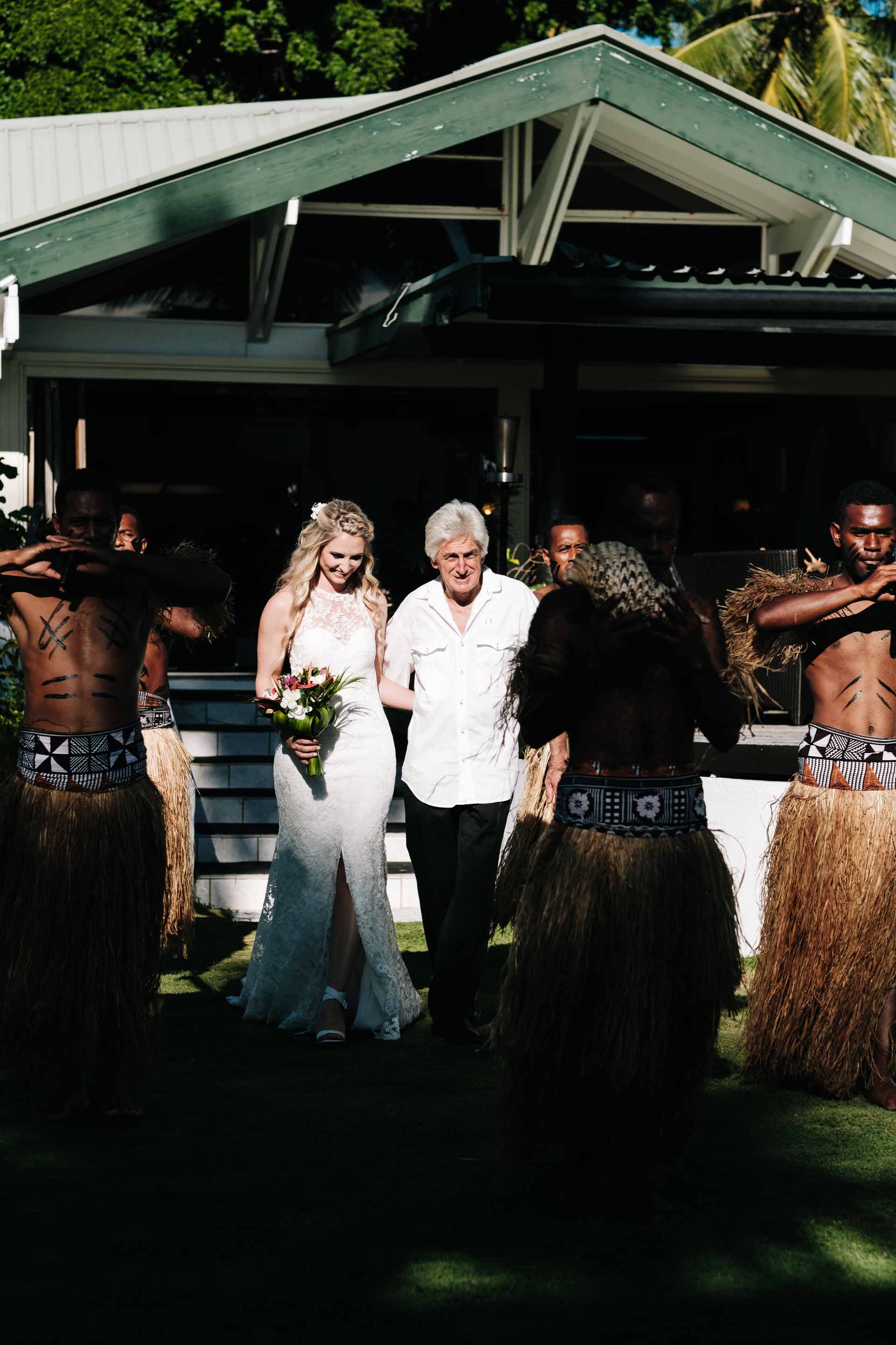 the bride and her father walking down the aisle escorted by Fijian warriors to the wedding arch