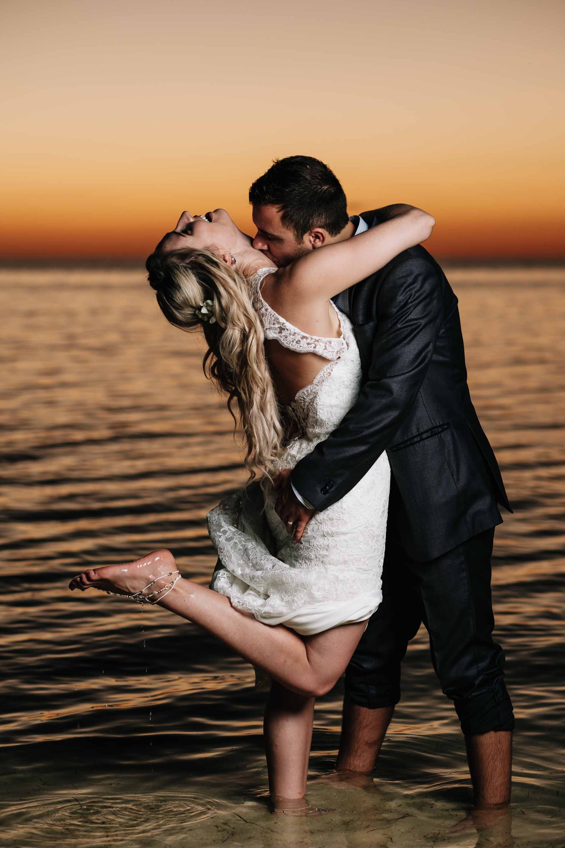 the bride and groom have fun in the warm water with the stunning backdrop of deep reds and oranges of a sunset