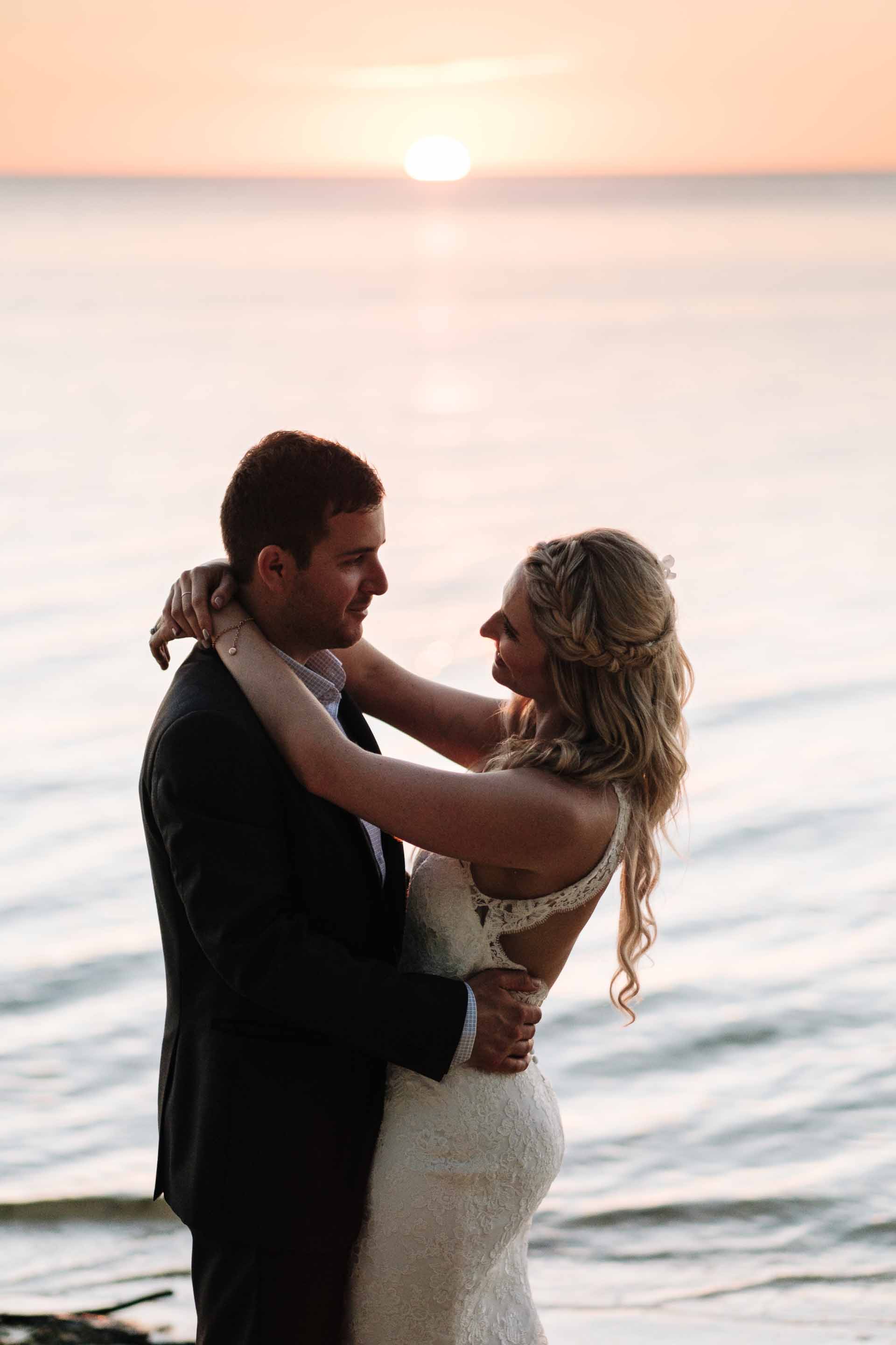 the bride and groom hold each other as the sun is about to dip behind the horizon
