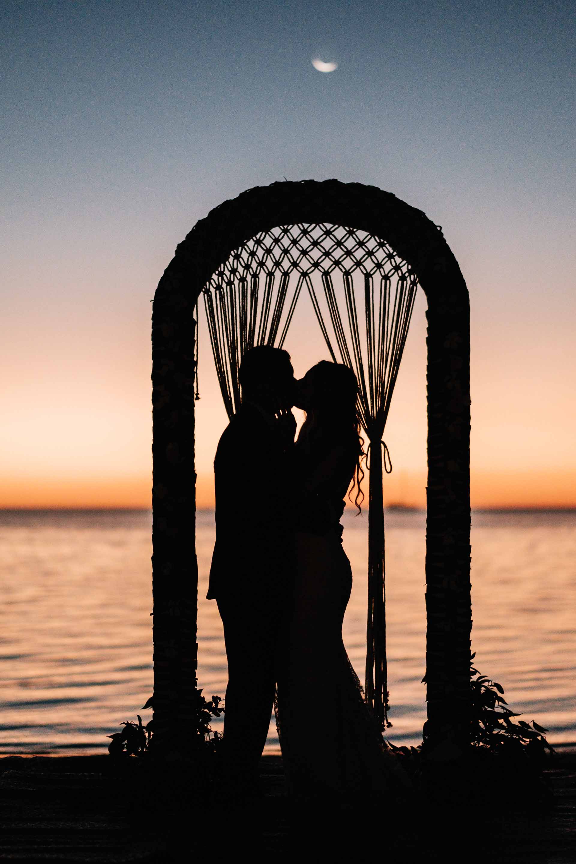 the bride &amp; groom share a kiss silhouetted against the calm warm seas and their macrame wedding arch just after the sun has set with the moon above them