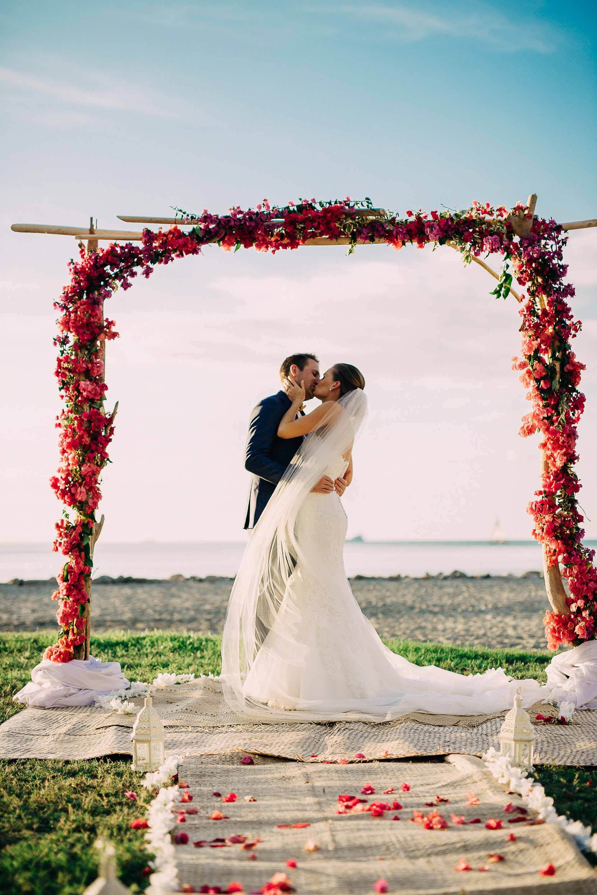 Fiji bride and groom kiss under their beautiful red bougainvillea and drift wood arch after their beach wedding.
