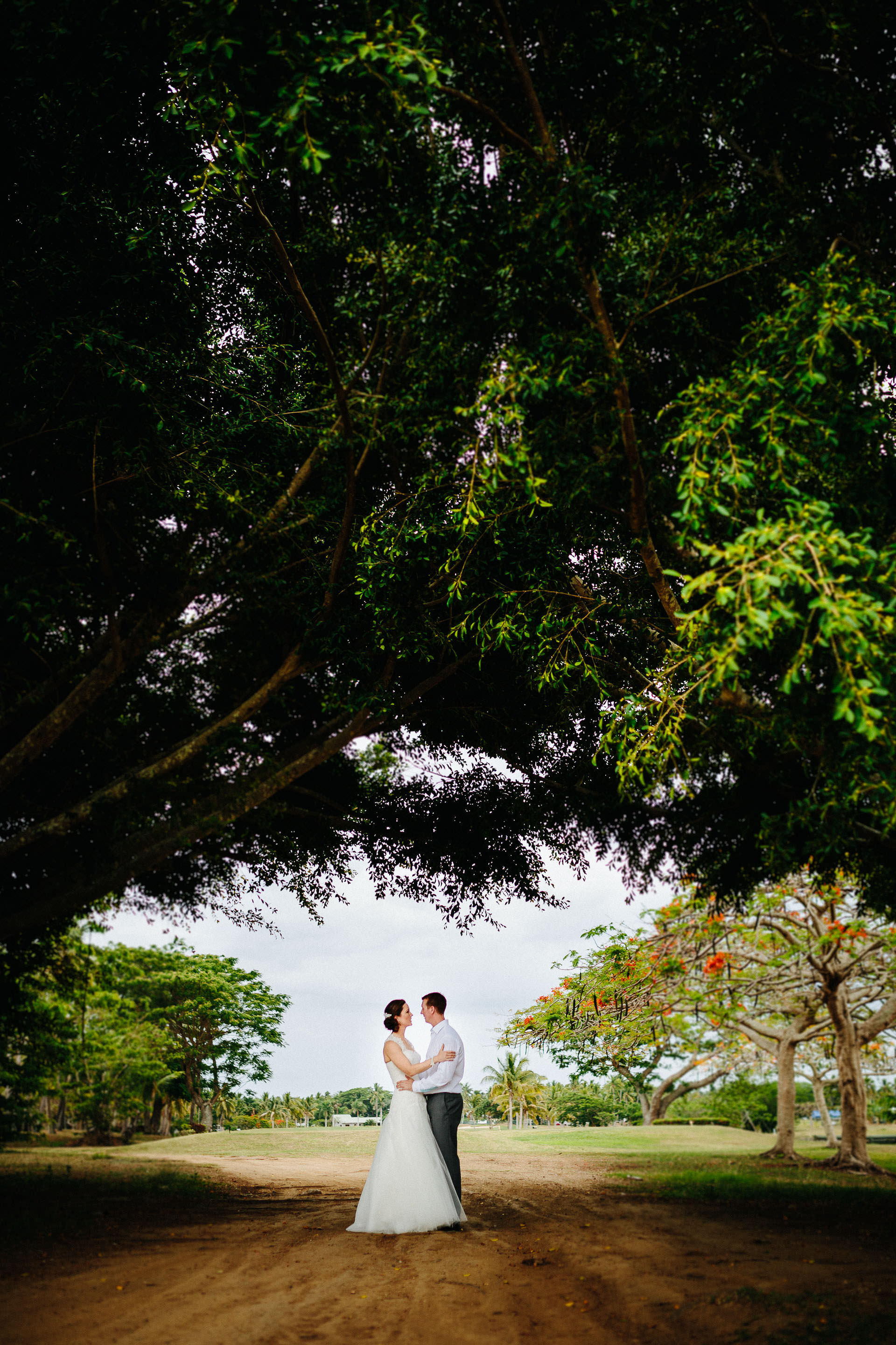 The bride and groom pose for a photo under two large trees