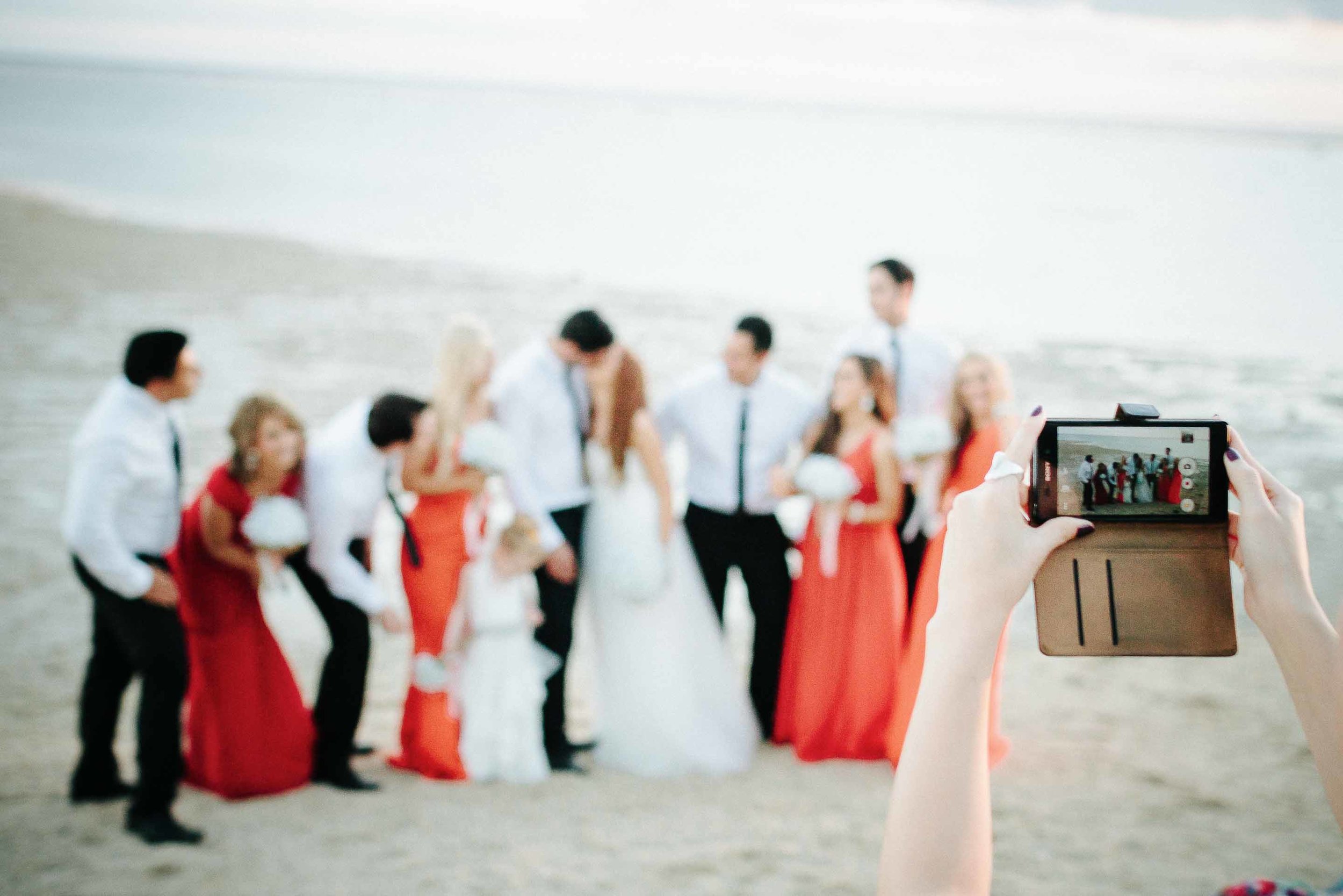 a guest taking a picture of the bridal party on the beach at sunset with their camera phone