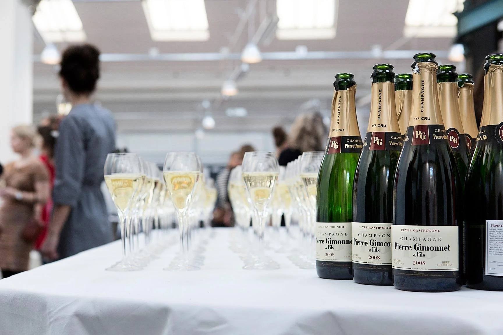 Flasker med champagne og flere færdigfyldte champagneglas på en hvid dug, med baggrund af folk til en fest.