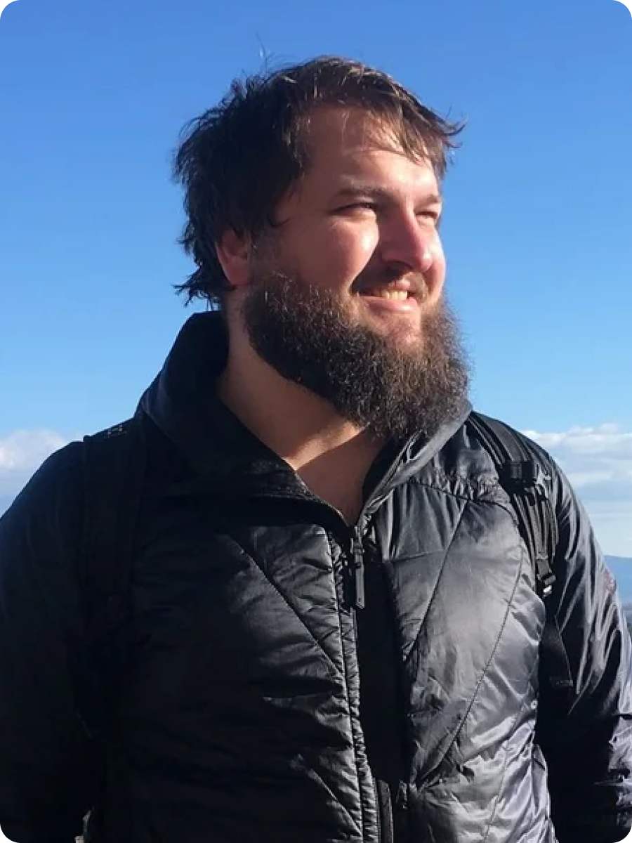 Mand iført sort jakke med skæg, står foran et landskab med marker og træer.