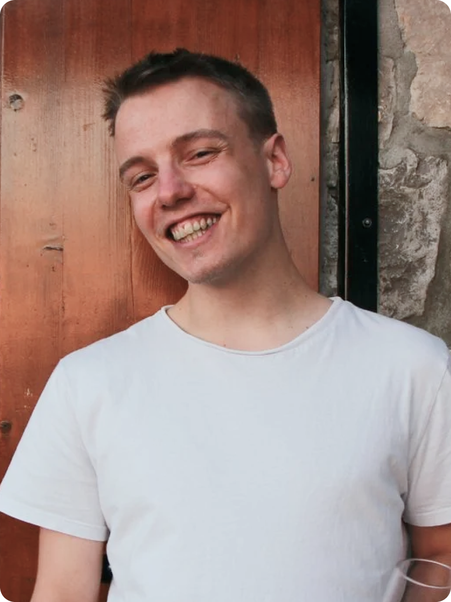 Ung mand i en hvid t-shirt står foran en stenvæg med en træluge og holder et glas rødvin.