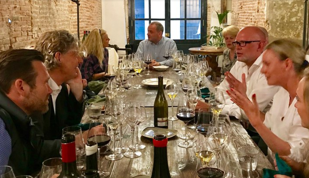 Mennesker sidder ved et langbord i en restaurant, der er dækket med vinflasker, glas, tallerkener og bestik.