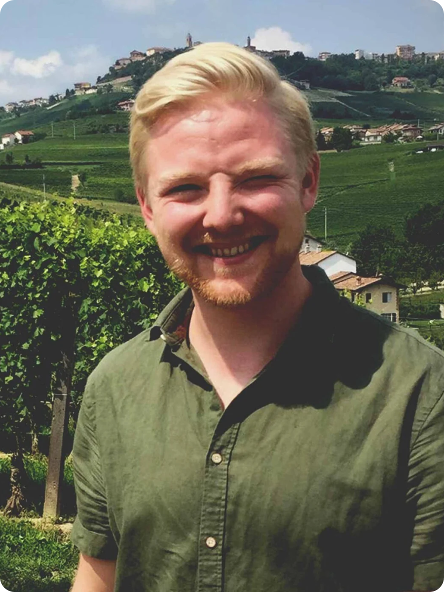 Mand i grøn skjorte smiler foran vinmarker med bakkede landskaber i baggrunden.
