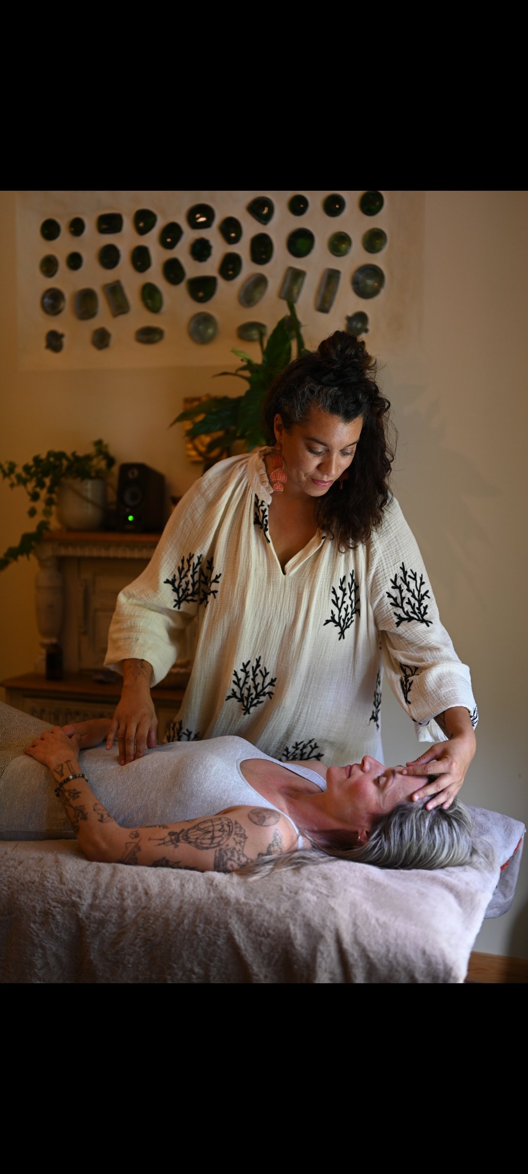 A woman with gray hair and tattoos lying on a massage table, receiving a head massage from a woman with curly dark hair in a room decorated with plants and artwork.