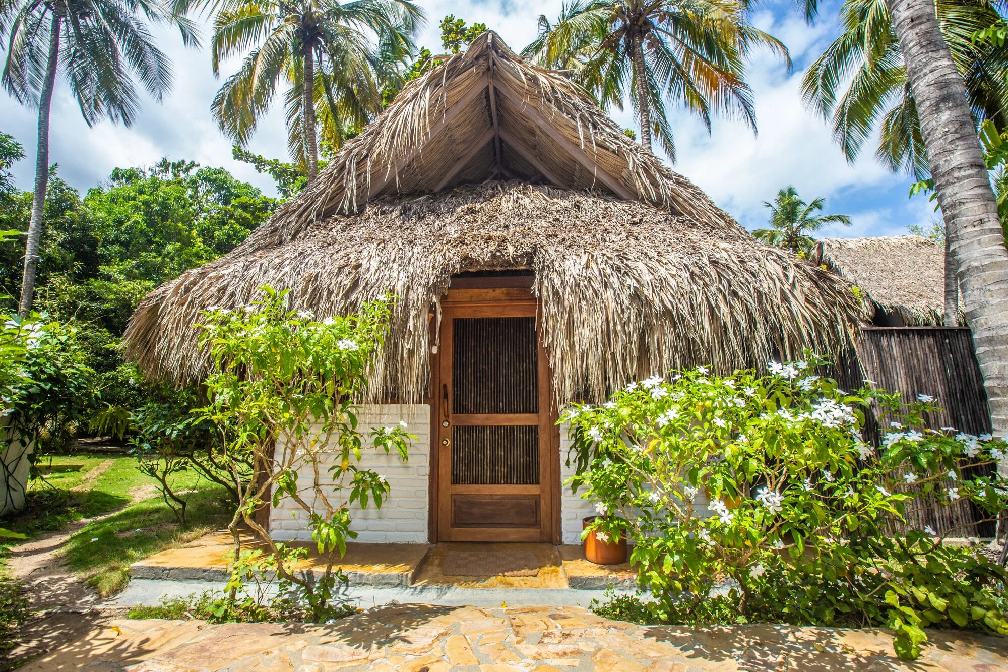 A tropical hut with a thatched roof, surrounded by lush greenery and flowering bushes, under a blue sky with some clouds.