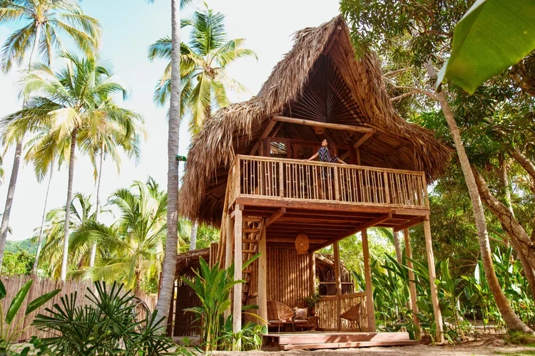 A two-story tropical treehouse with a thatched roof, wooden balcony, and outdoor seating, surrounded by lush green palm trees and foliage.