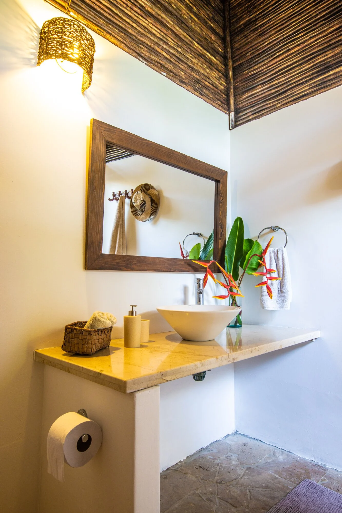 A bathroom with a white vessel sink on a marble countertop, decorated with a colorful flower arrangement and a plant, with a round mirror, a wooden wall-mounted frame, a wicker basket, soap dispenser, towel, and a hat on the wall, illuminated by a woven wall sconce light.